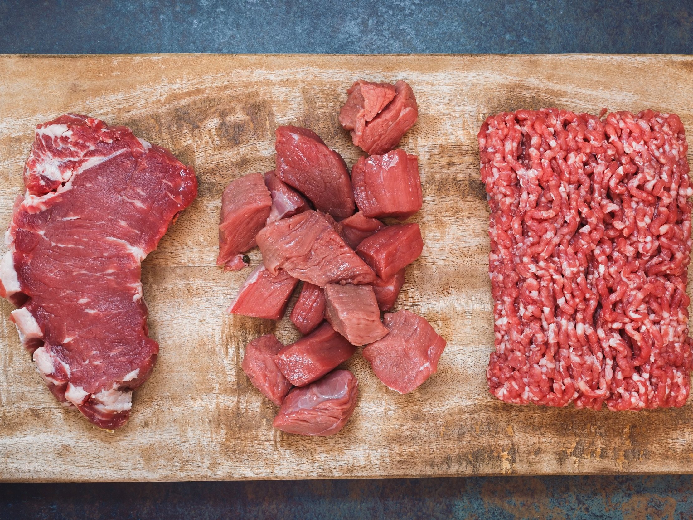 Raw beef cuts including a ribeye steak, cubed beef, and ground beef displayed on a wooden cutting board, highlighting different meat preparations for cooking.