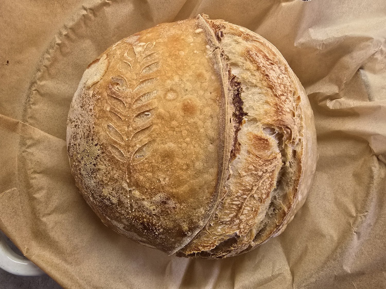 Artisan sourdough bread with a decorative leaf pattern, resting on parchment paper. The crust is golden brown and textured, showcasing the bread's rustic appearance and freshly baked quality.